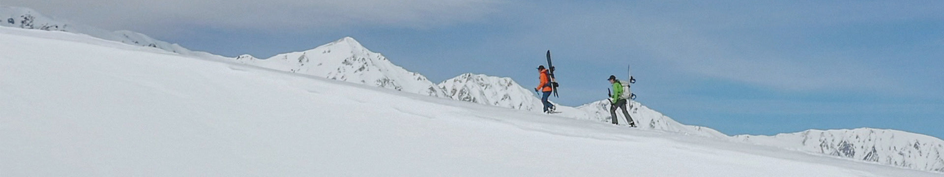 Two backcountry skiers snowshoeing uphill with skis strapped to their packs on a snowy alpine ridge.