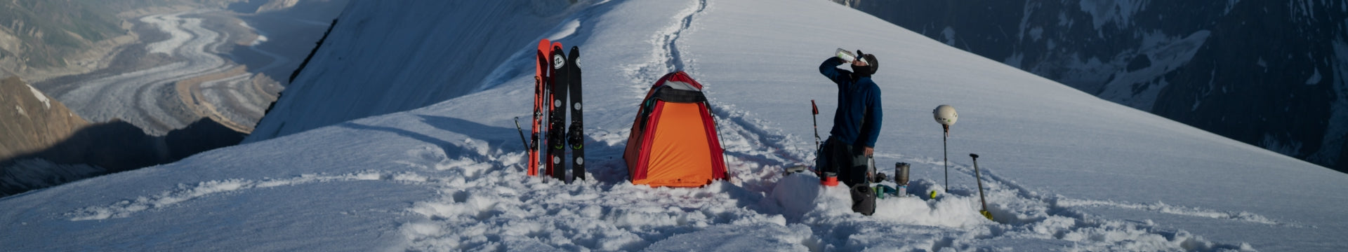 Winter camper on a snowy ridge with a tent and skis.
